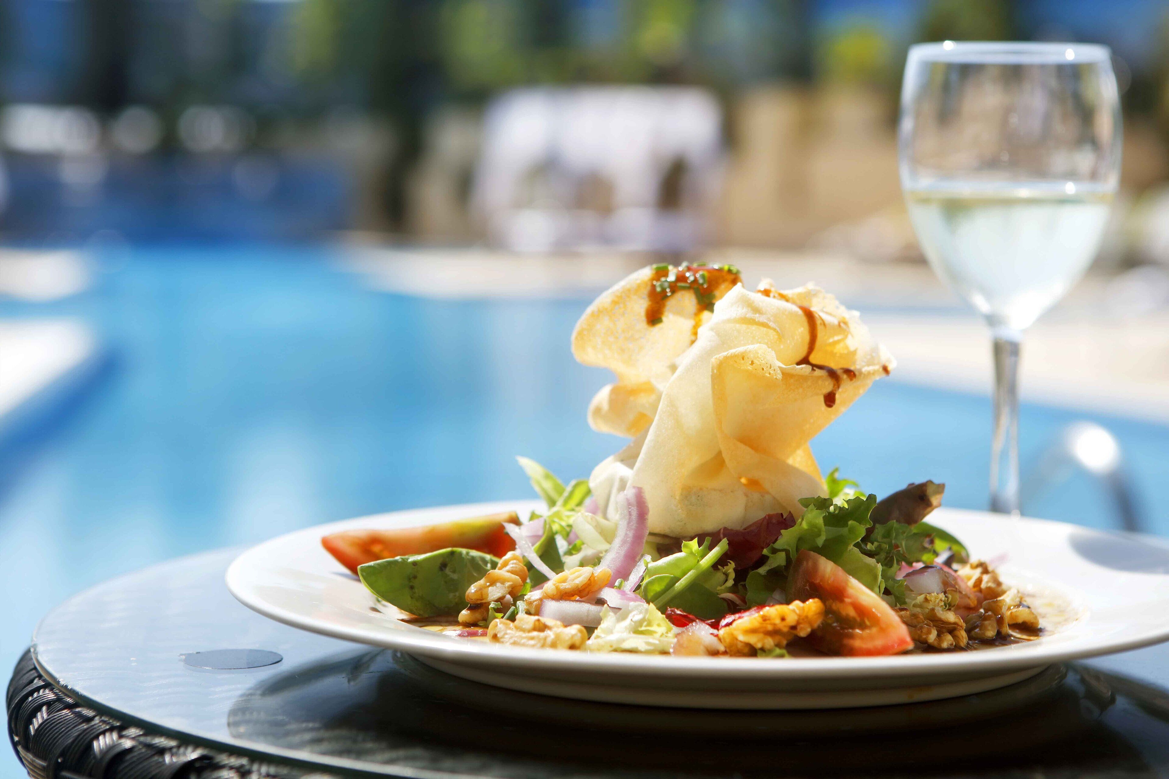 Une assiette de nourriture, avec une salade, et un verre de vin blanc, sur une table près d’une piscine.
