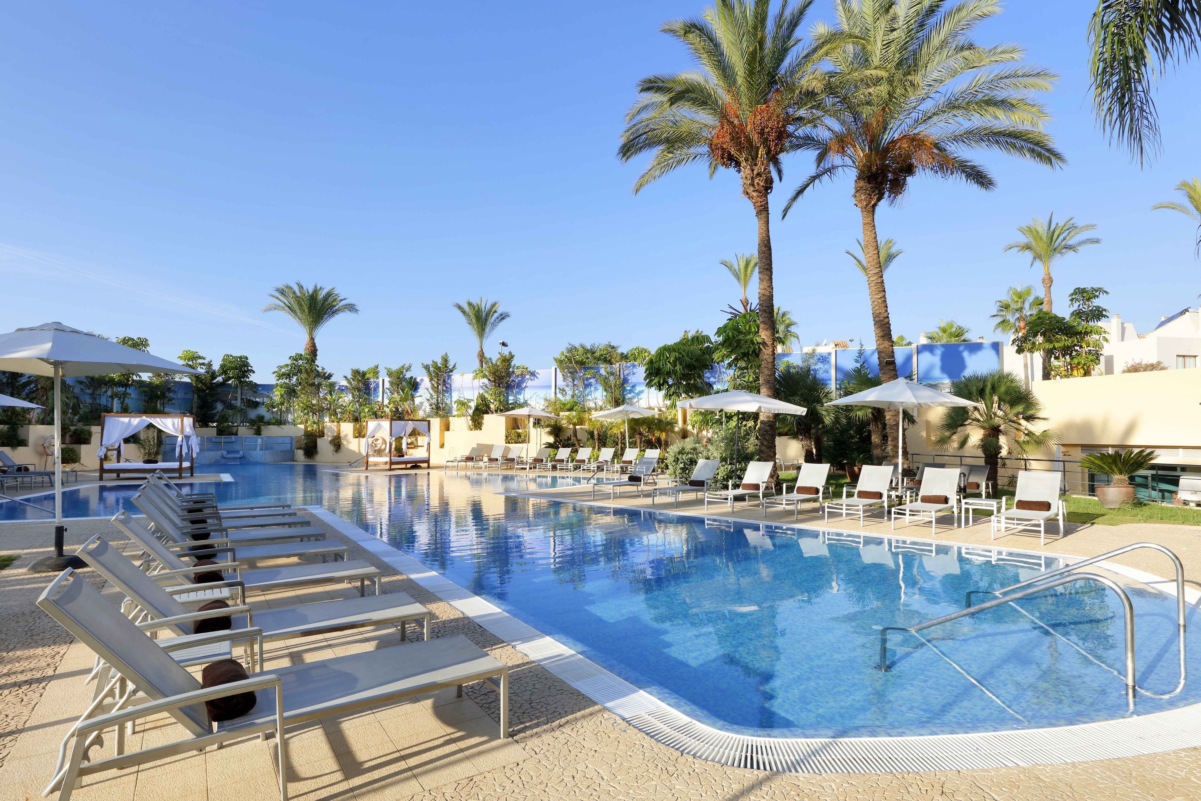 A pool area with lounge chairs and umbrellas, surrounded by palm trees and a clear sky.