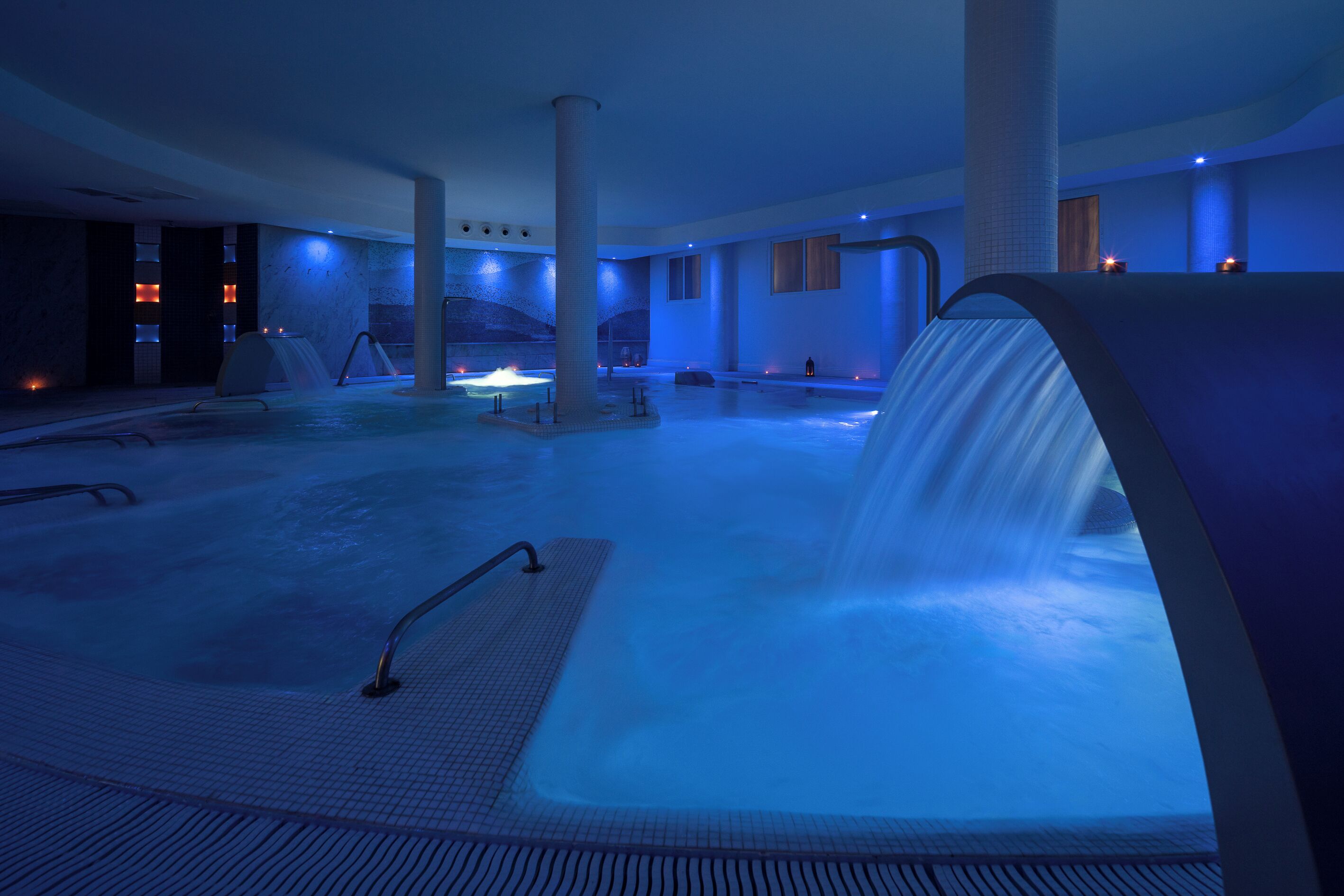 Amplia zona interior con bañera de hidromasaje y una cascada, rodeada de pilares y una iluminación tenue.