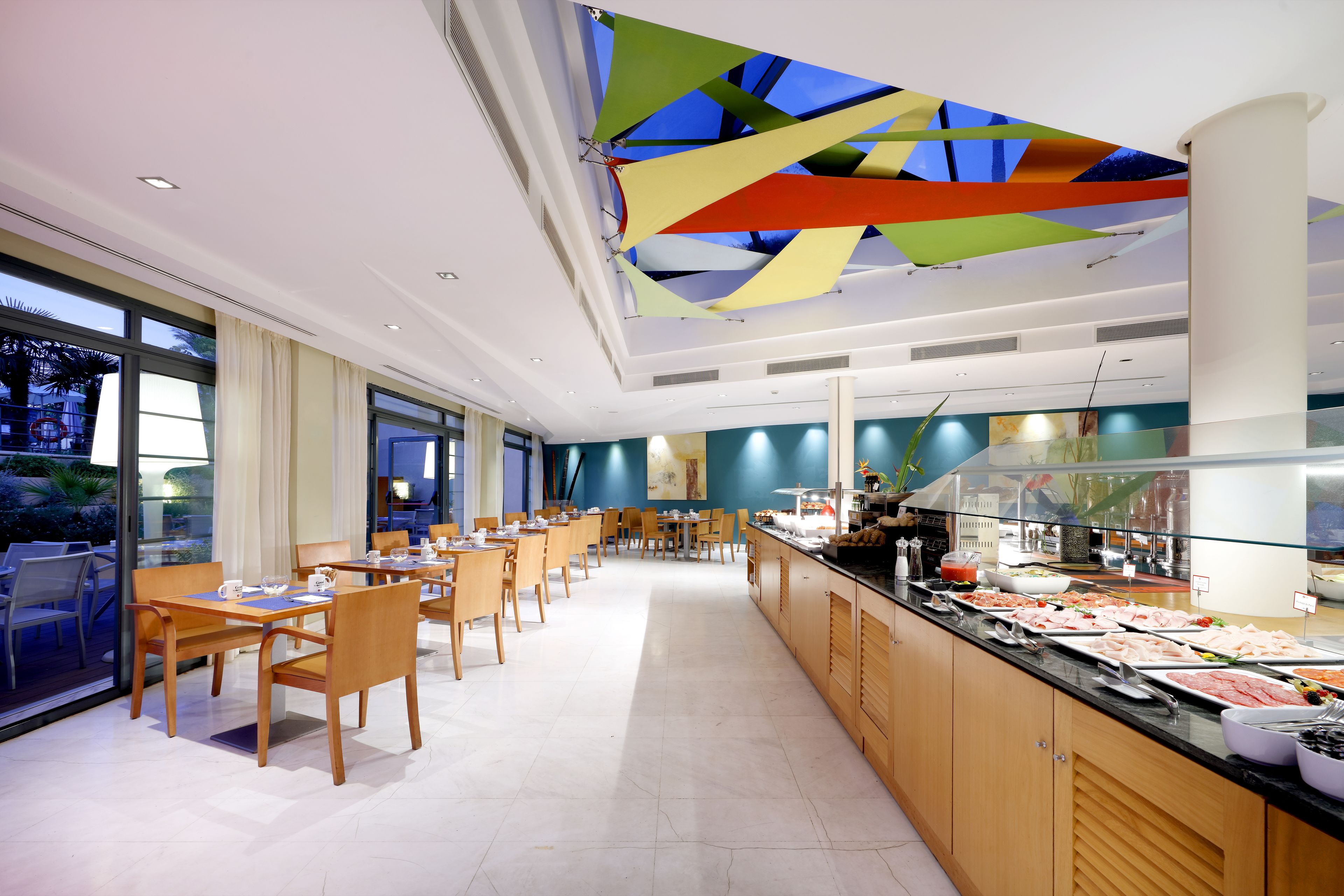 Un espace de restauration moderne avec de longues tables et des chaises, un buffet proposant une variété de plats, et un plafond orné de motifs géométriques colorés.