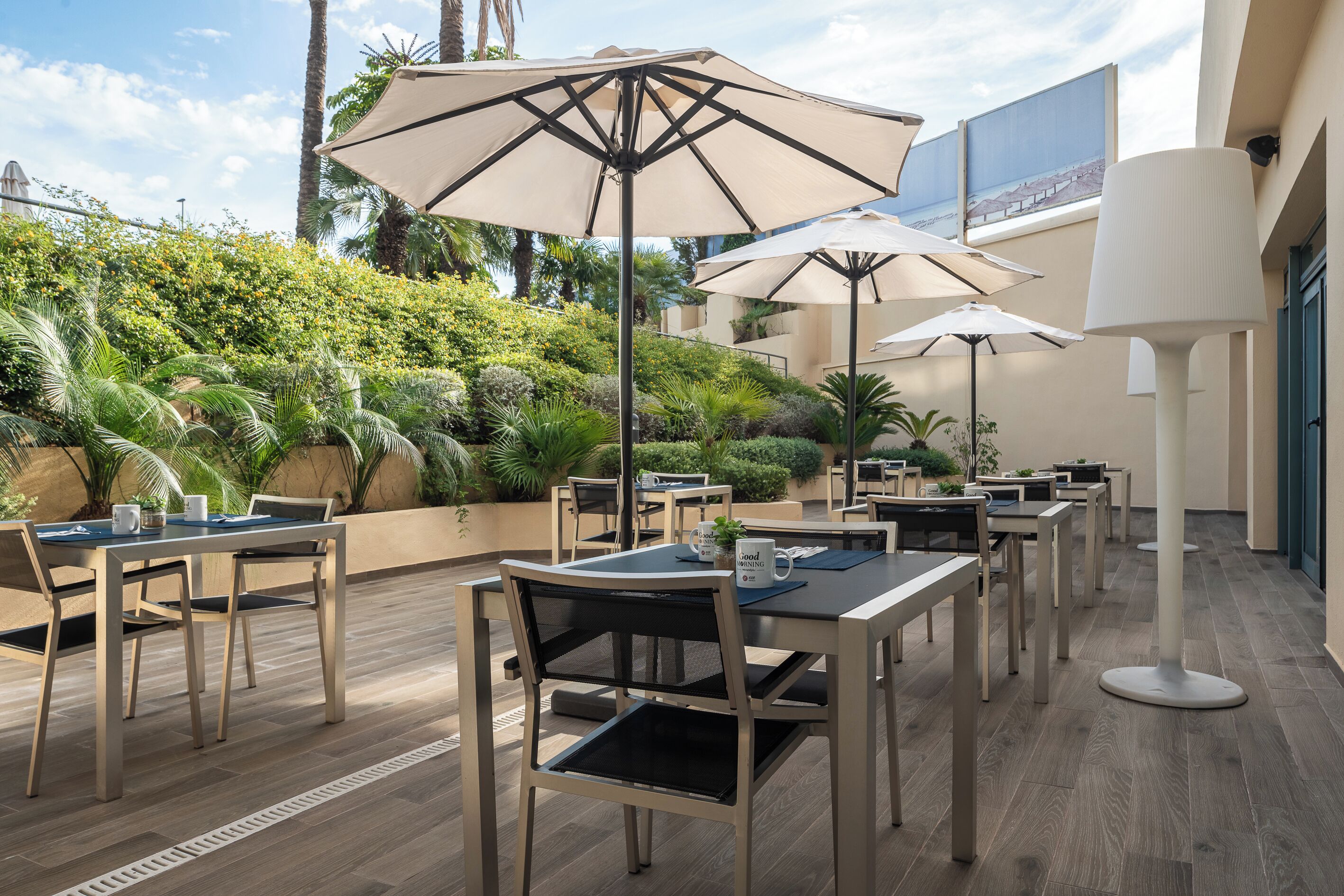 Un área de comedor al aire libre con mesas, sillas y sombrillas, rodeada de abundante follaje y palmeras.