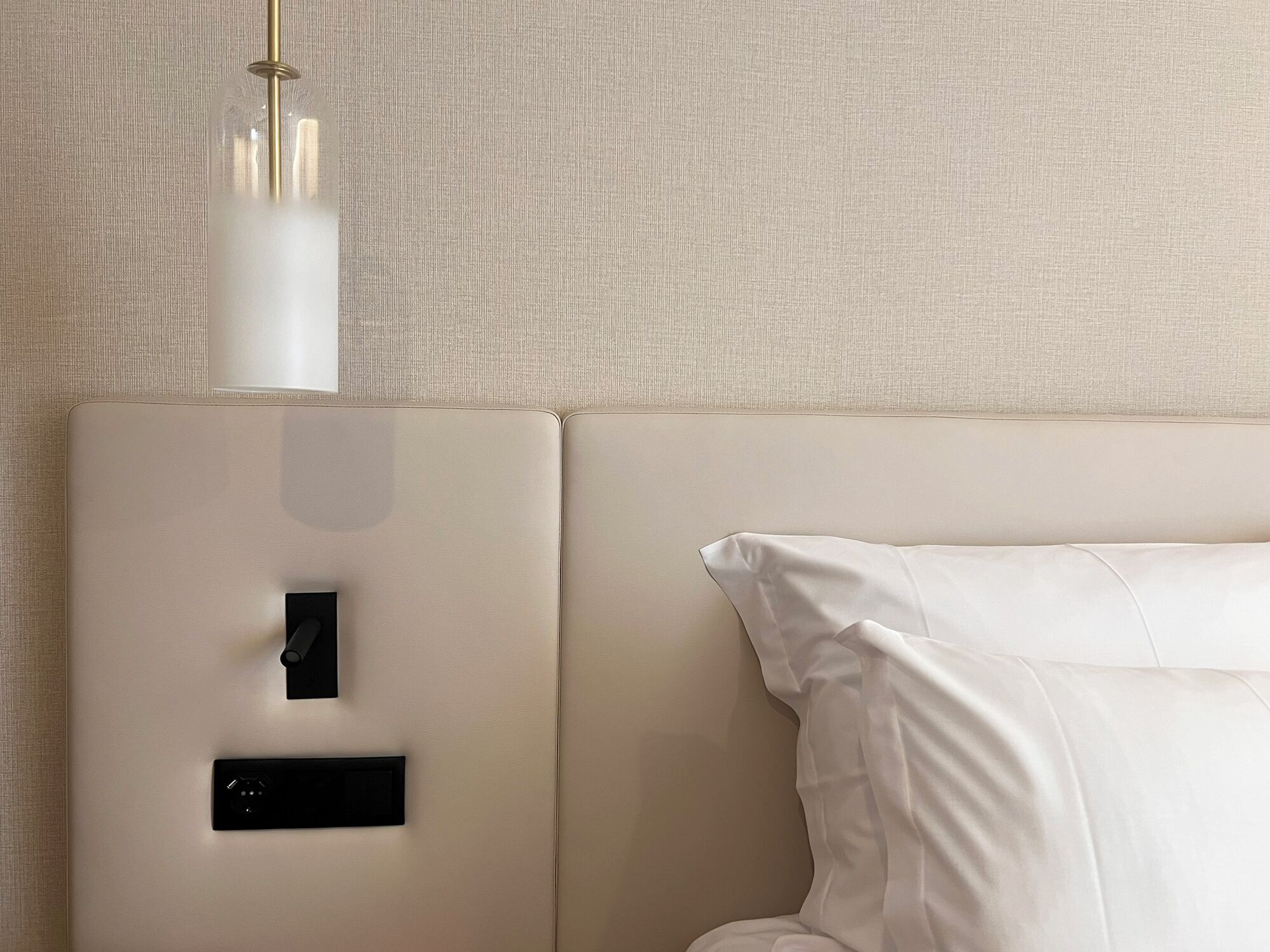 A modern hotel room with a minimalist design, featuring a bed with white linens, a control panel for room amenities, and a pendant light.