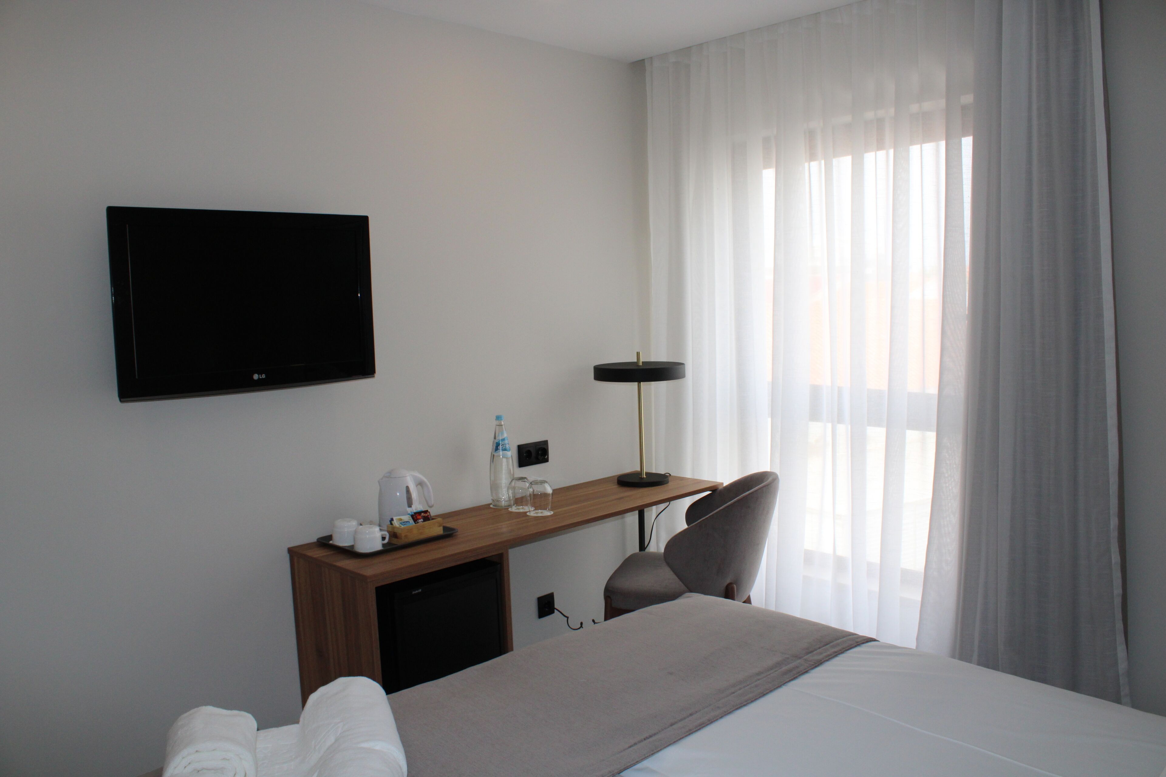A hotel room with a bed, a desk with a flat-screen TV, a chair, a desk lamp, and a window with sheer curtains.