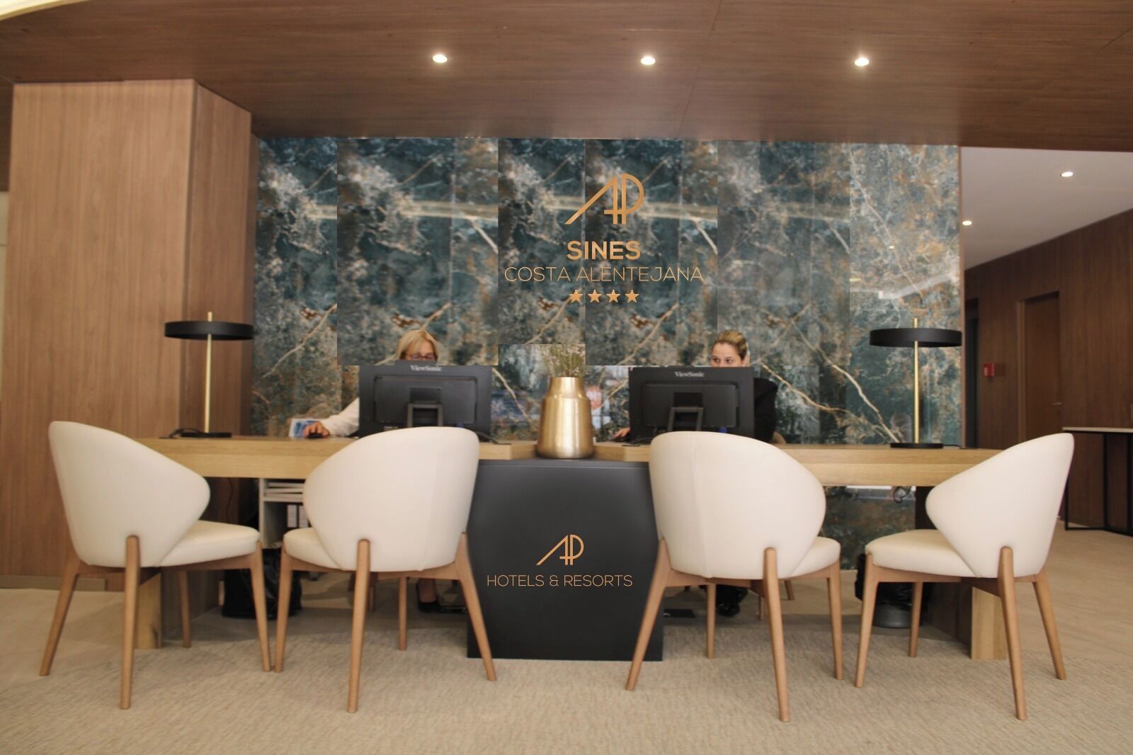 Una recepción con dos empleados, una pared de mármol y el logotipo de 'COSTA ALENTEJANA HOTELS & RESORTS'.
