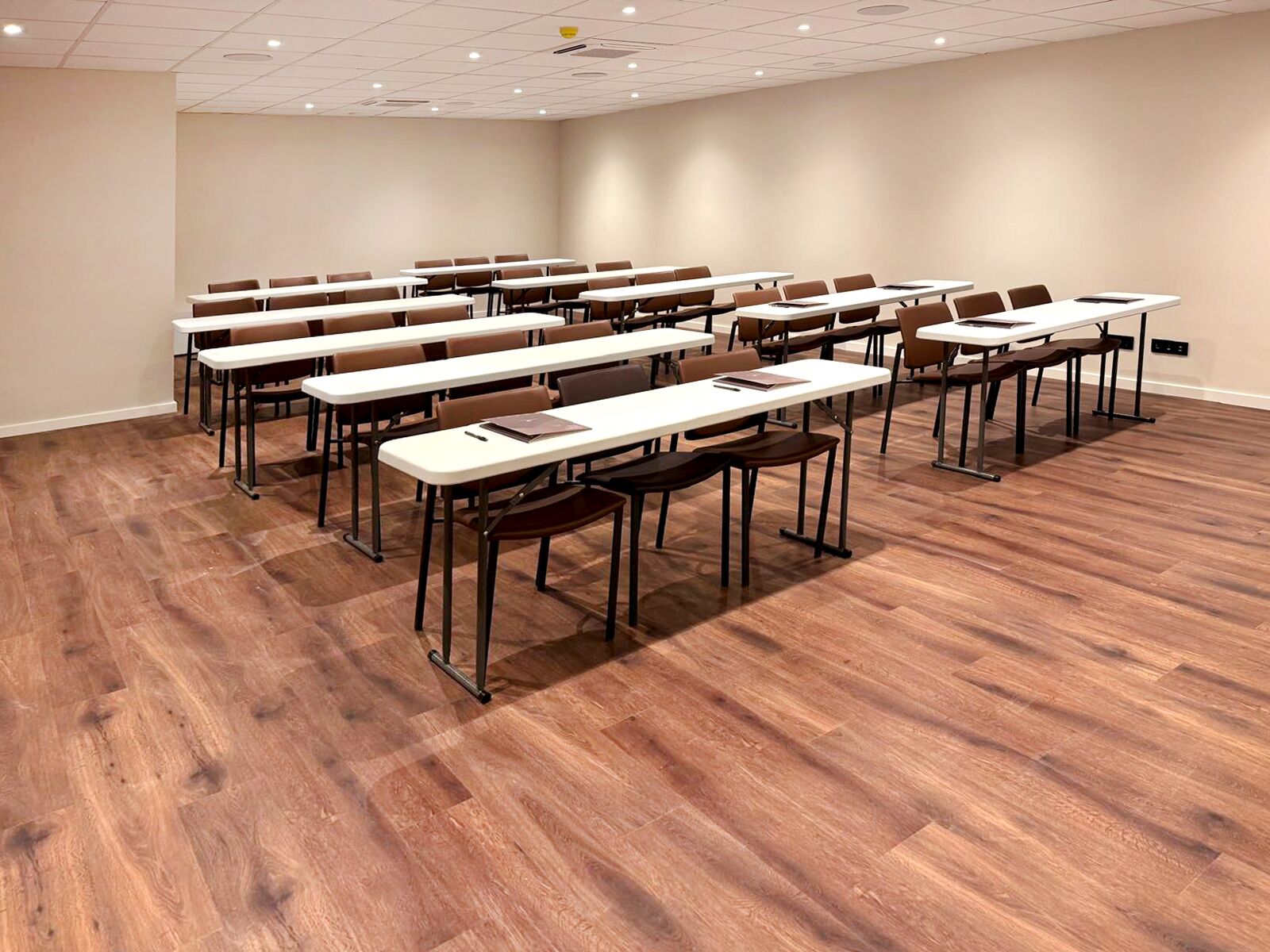 A conference room with multiple tables and chairs, wooden flooring, and a plain wall.