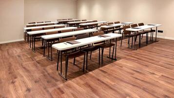 A conference room with multiple tables and chairs, wooden flooring, and a plain wall.