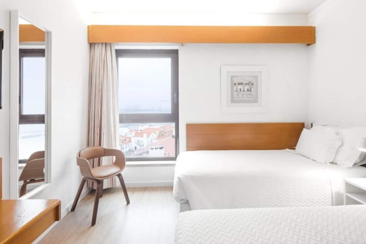 Quarto de hotel com cama, poltrona, escrivaninha, janela com vista e quadro na parede.