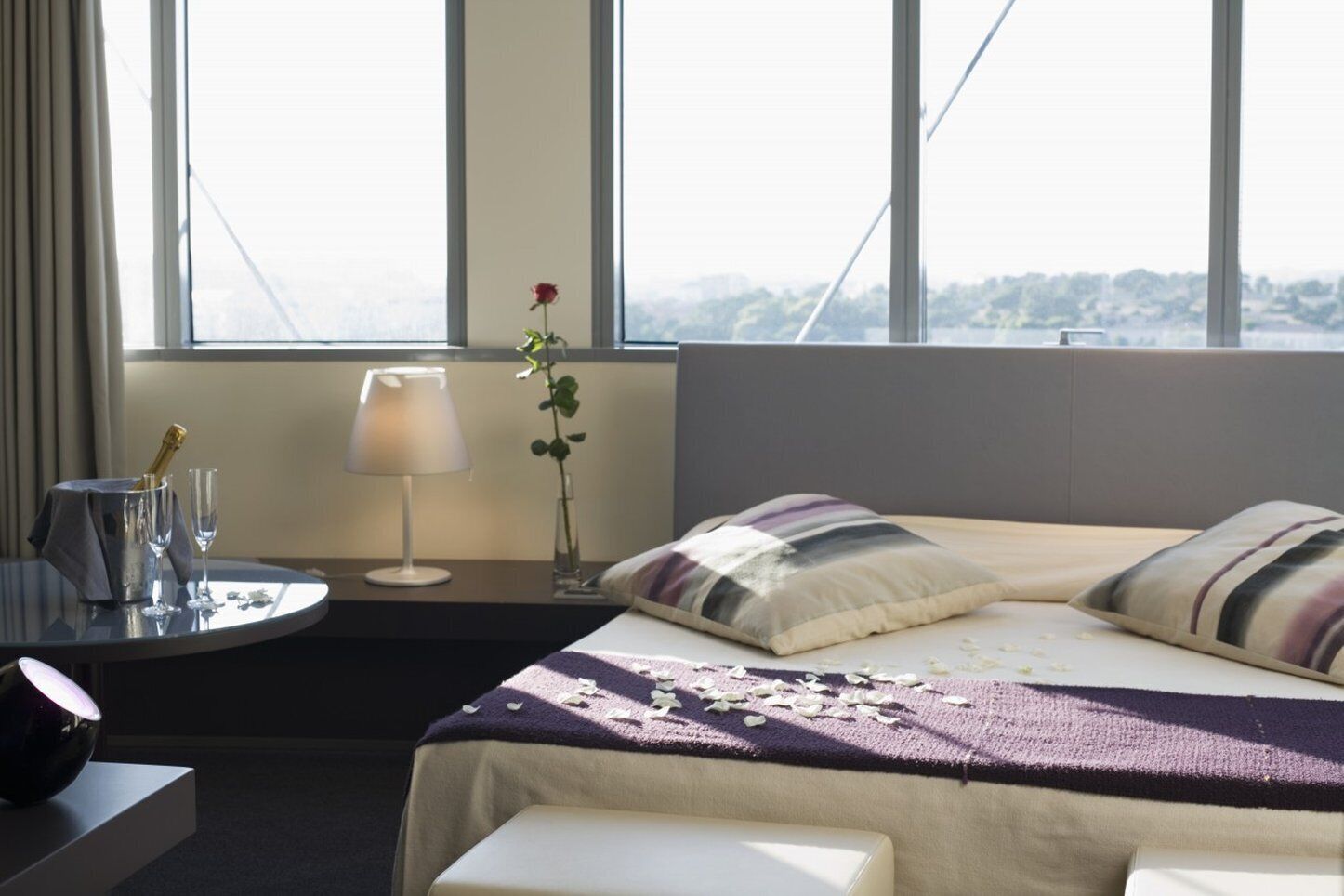 Habitación de hotel con cama, mesita de noche, lámpara y vistas a la ciudad.