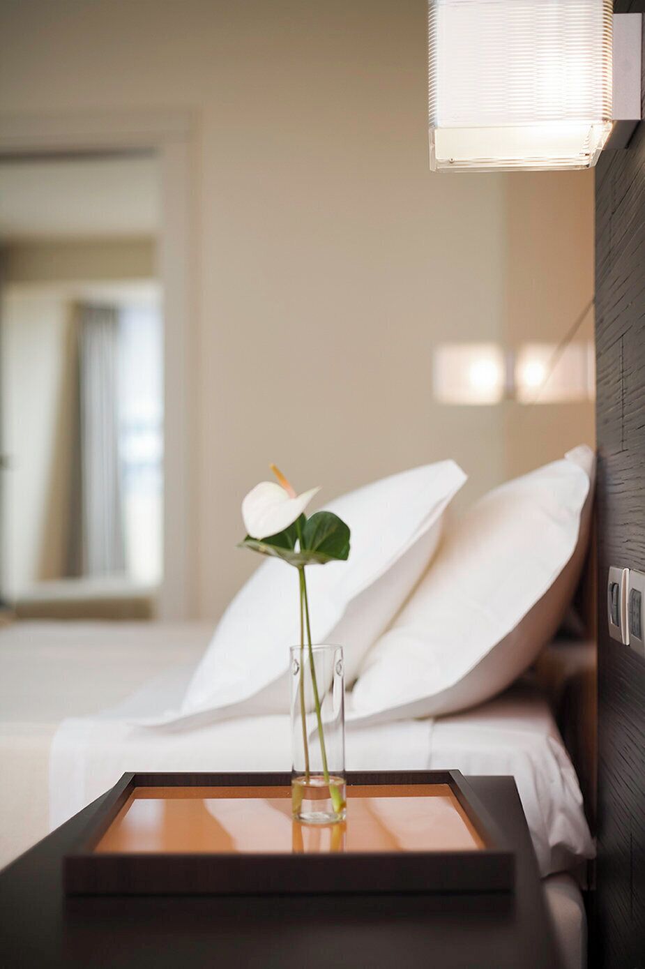 A hotel room with a bed, a bedside table with a vase of flowers, and a wall-mounted light fixture.