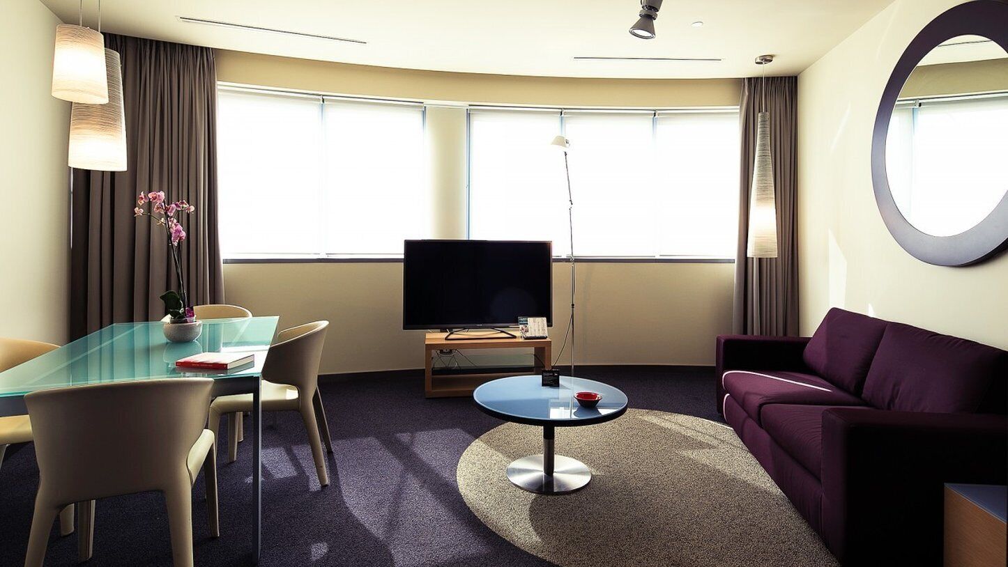 A hotel room with a glass table, a flat-screen TV, a purple sofa, and a large mirror.