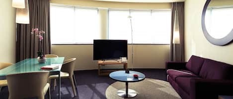 A hotel room with a glass table, a flat-screen TV, a purple sofa, and a large mirror.