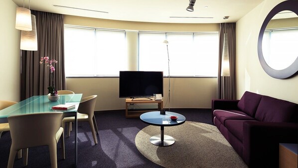 A hotel room with a glass table, a flat-screen TV, a purple sofa, and a large mirror. - UNA HOTELS T Hotel Cagliari (Cagliari)