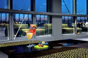 Una mesa con una copa de cóctel rojo, una copa de cóctel amarillo y un pequeño plato con comida, frente a amplias ventanas con vistas a un paisaje urbano.