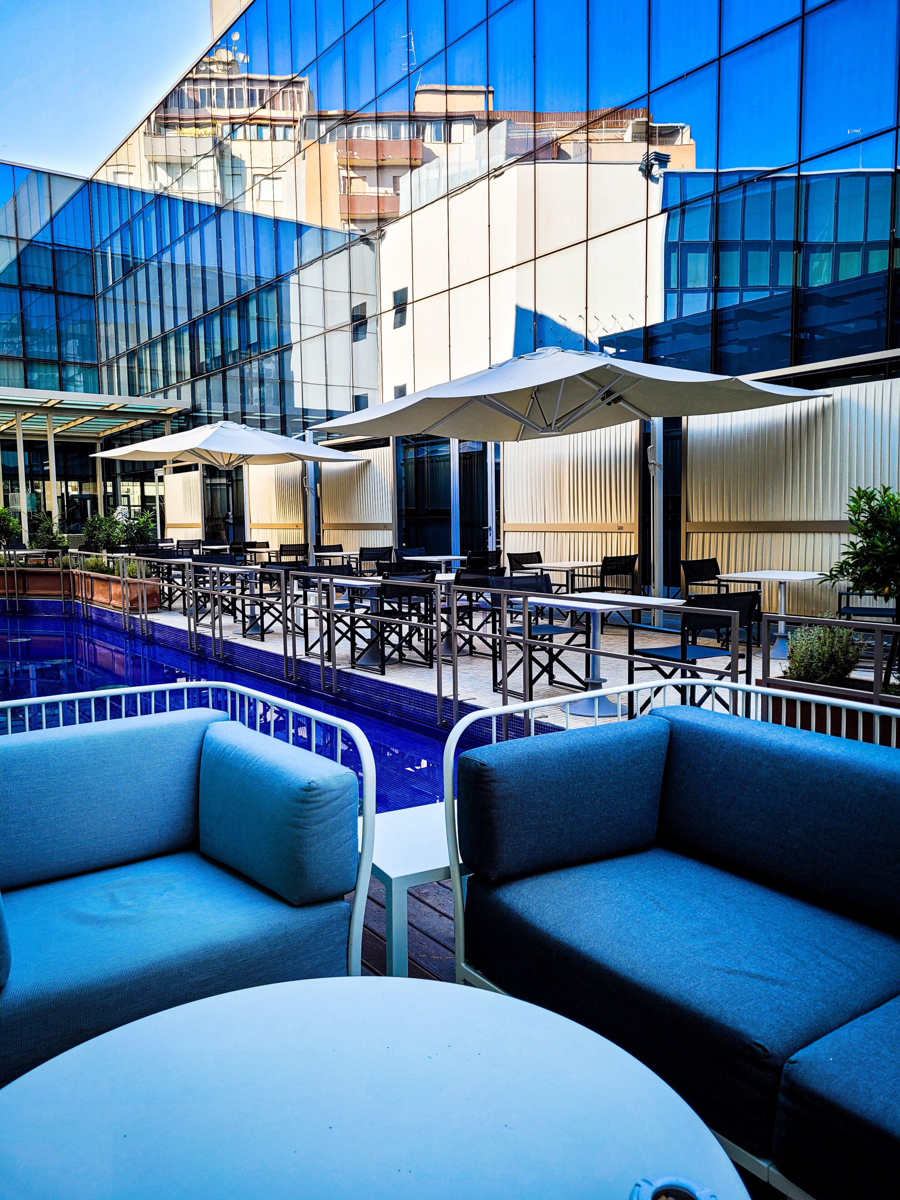An outdoor seating area with tables and chairs, a pool, and a modern building with reflective glass windows.