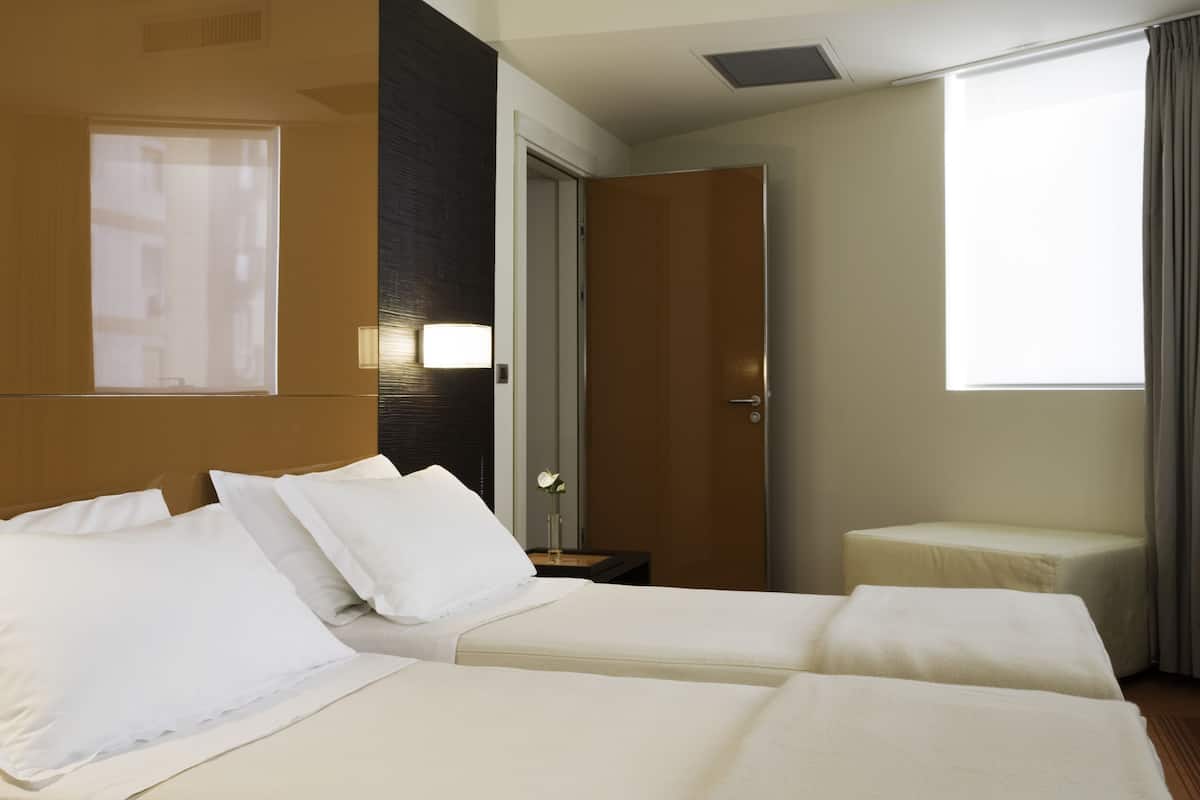 A hotel room with two beds, a nightstand, a lamp, a window, and a door.