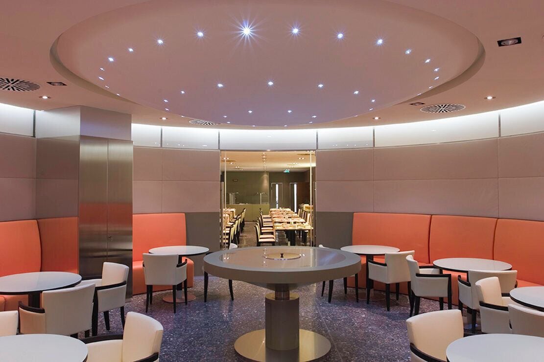 A modern interior with round tables and orange seating, a central light fixture, and a view into a dining area.