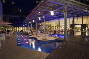 Zona de piscina interior con asientos y mesas, rodeada por paredes de vidrio y un paseo cubierto.