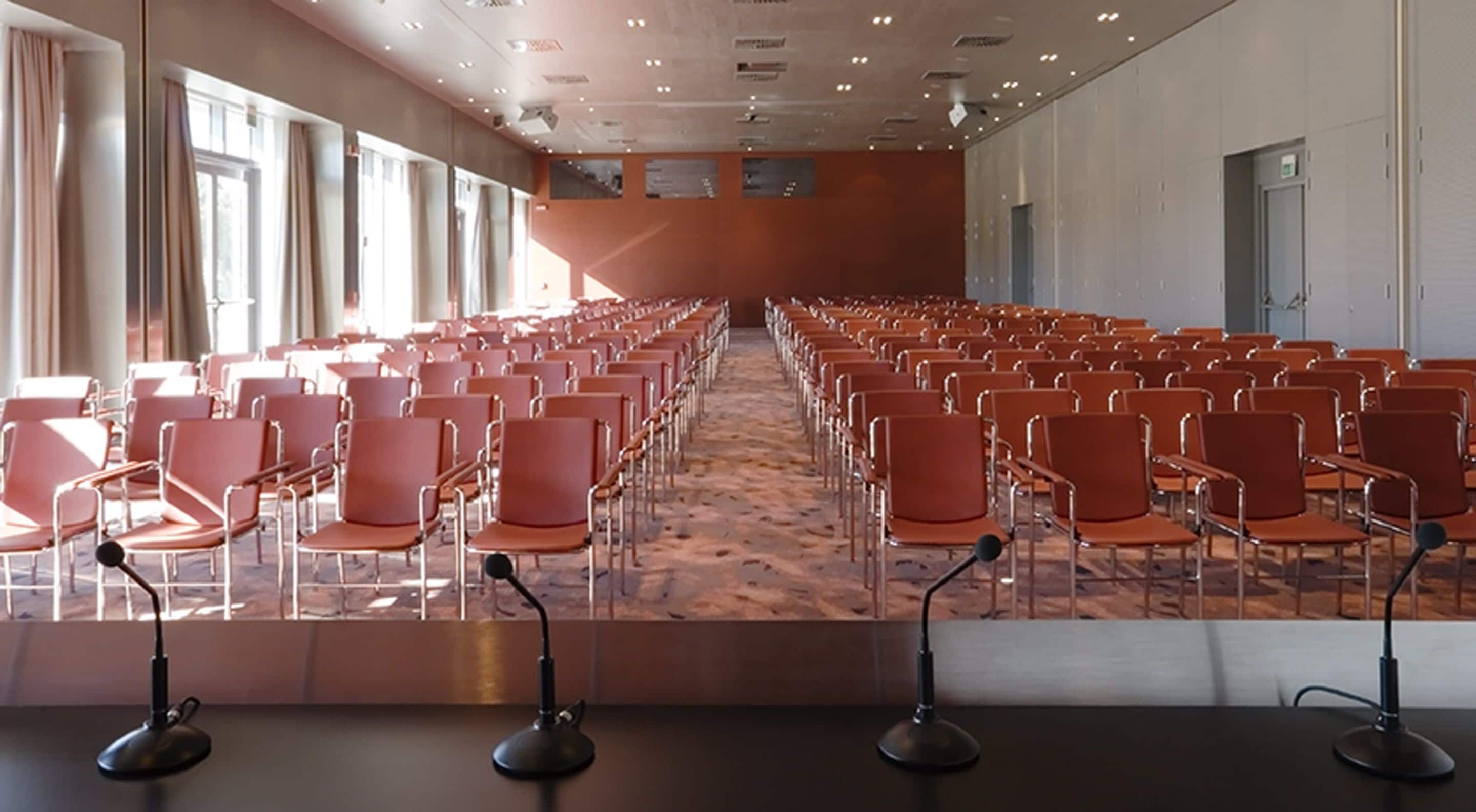 A conference room with rows of orange chairs, a carpeted floor, and a long table at the front with microphones.