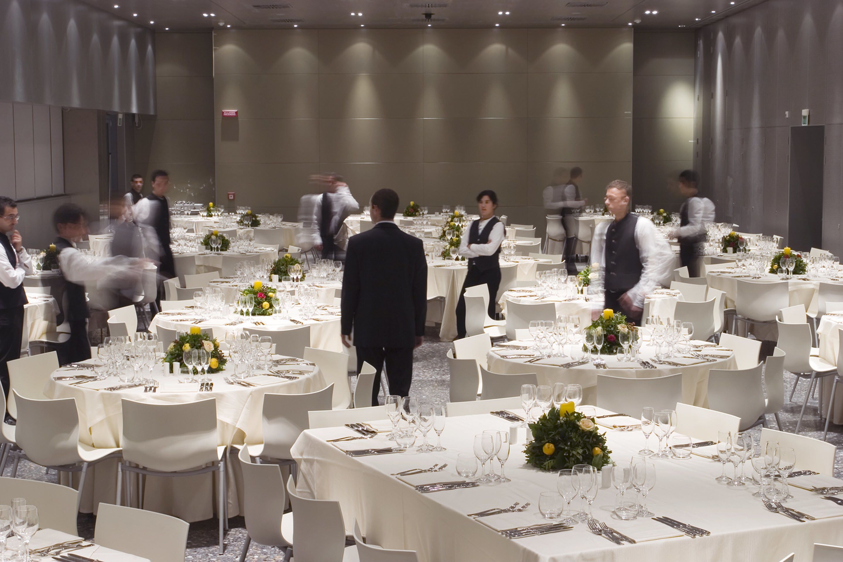 Un salón de banquetes con mesas redondas dispuestas para cenar, cada una con manteles blancos, sillas y cristalería.