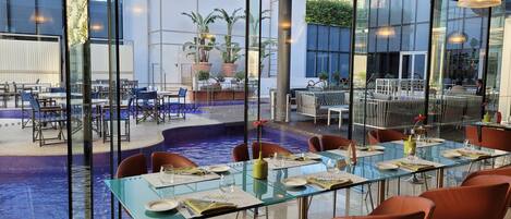 A modern indoor dining area with glass tables and chairs, a pool, and potted plants.