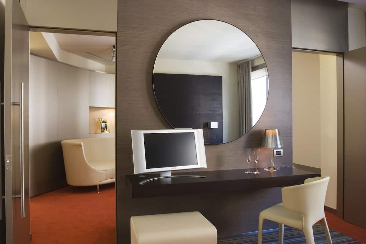 A modern hotel room with a round mirror, a flat-screen TV, a desk with a lamp, a chair, and a sofa.