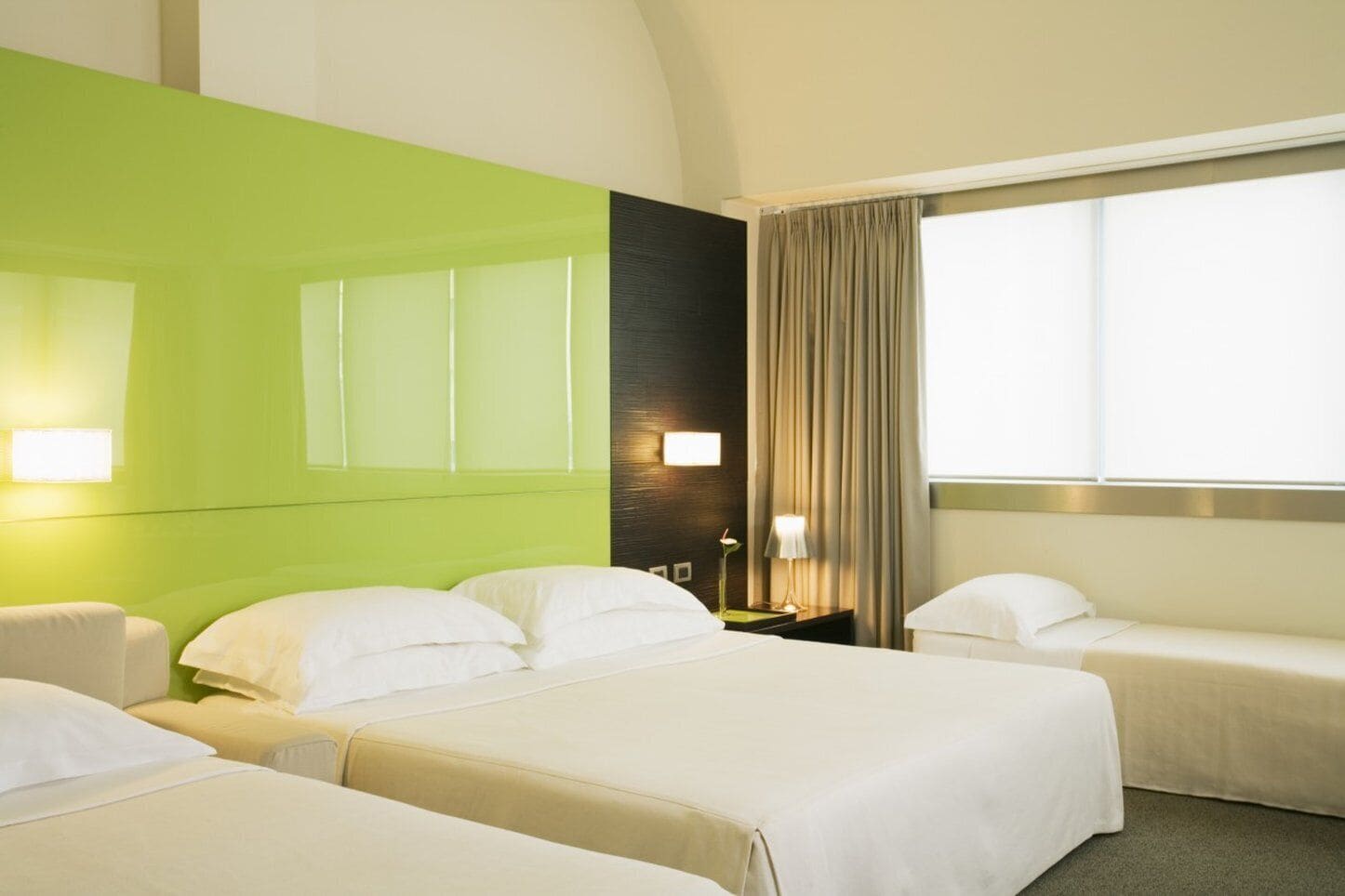 Habitación de hotel con dos camas, una pared de acento verde, un ventanal con cortinas y lámparas de noche.