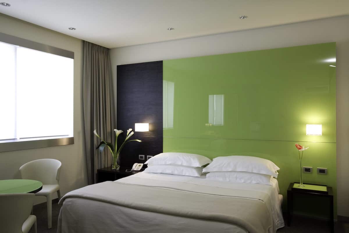 A hotel room with a bed, a chair, a nightstand, and a window with curtains.