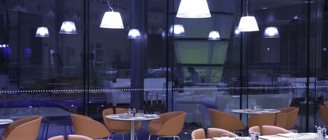 A modern dining area with round tables and orange chairs, large windows, and pendant lighting.