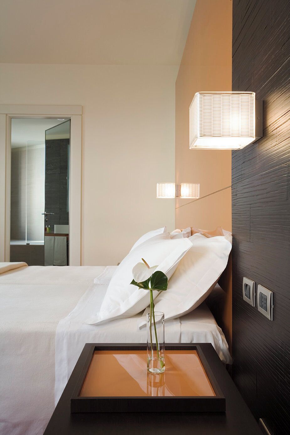 Una camera d'albergo con un letto, un comodino con un vaso, una lampada a parete e uno specchio.
