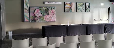 Un salón de conferencias con filas de sillas, un cuadro grande con motivos florales y un detector de incendios en el techo.