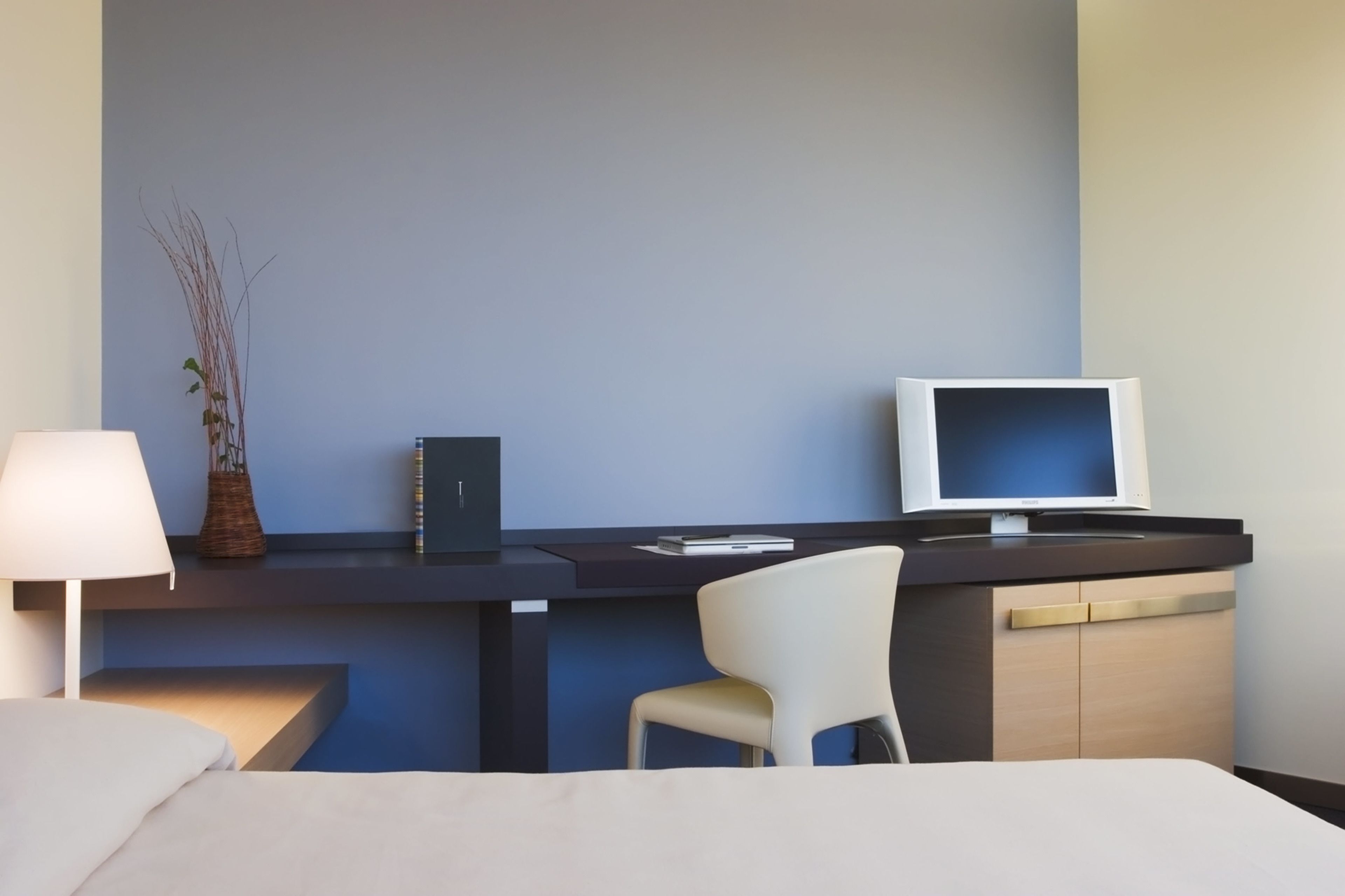 A hotel room with a bed, a desk with a computer monitor, a chair, and a lamp.