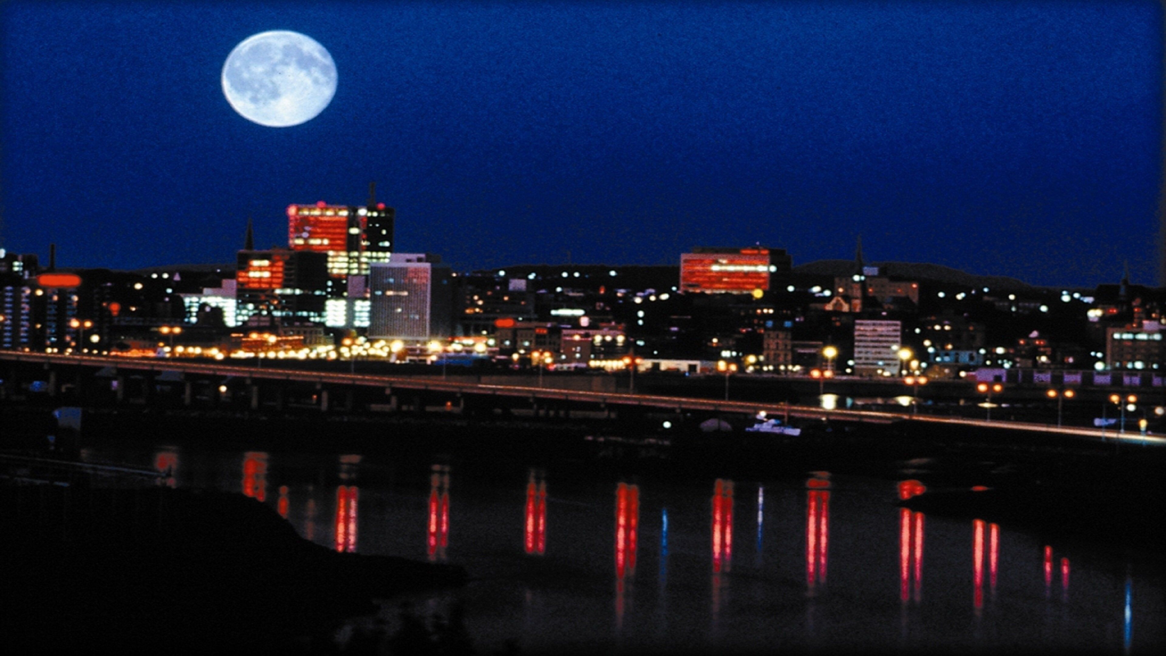 Une vue nocturne d’une ville, avec une pleine lune bien visible dans le ciel, des bâtiments illuminés et un pont qui reflète les lumières.