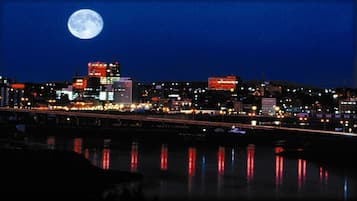 Une vue nocturne d’une ville, avec une pleine lune bien visible dans le ciel, des bâtiments illuminés et un pont qui reflète les lumières.