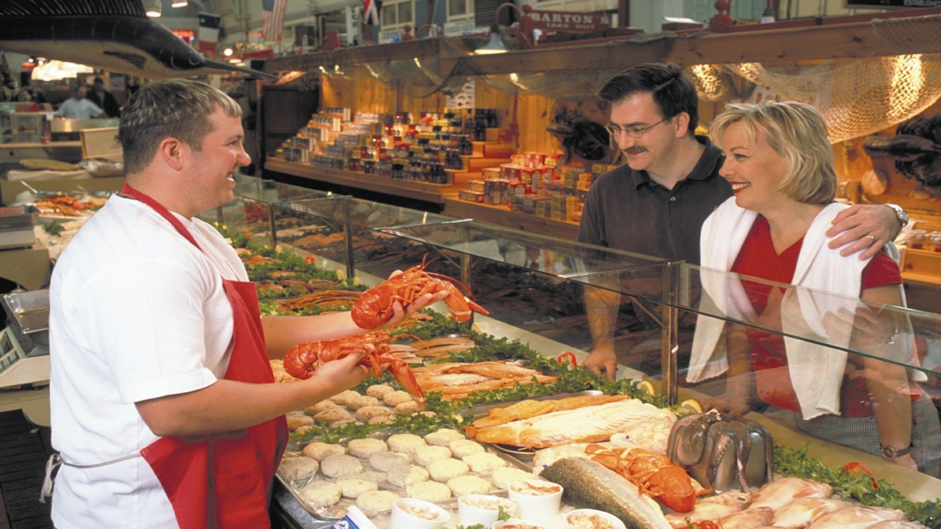 Una exhibición de mariscos con varios productos como langostas y pescado, una persona con delantal rojo y una barrera de vidrio.