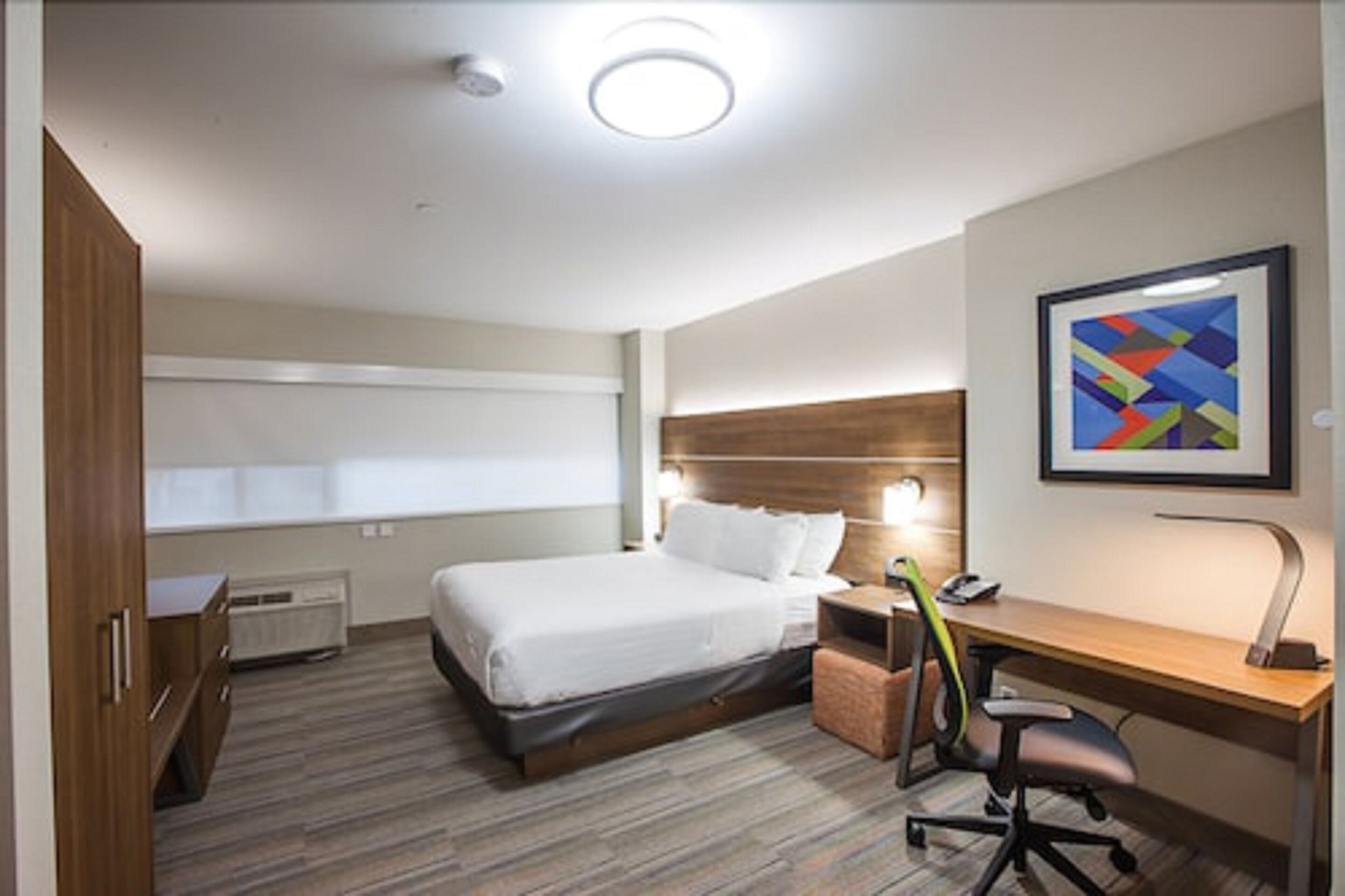 Ein Hotelzimmer mit Bett, Nachttisch, Schreibtisch, Stuhl und einem gerahmten abstrakten Gemälde an der Wand.