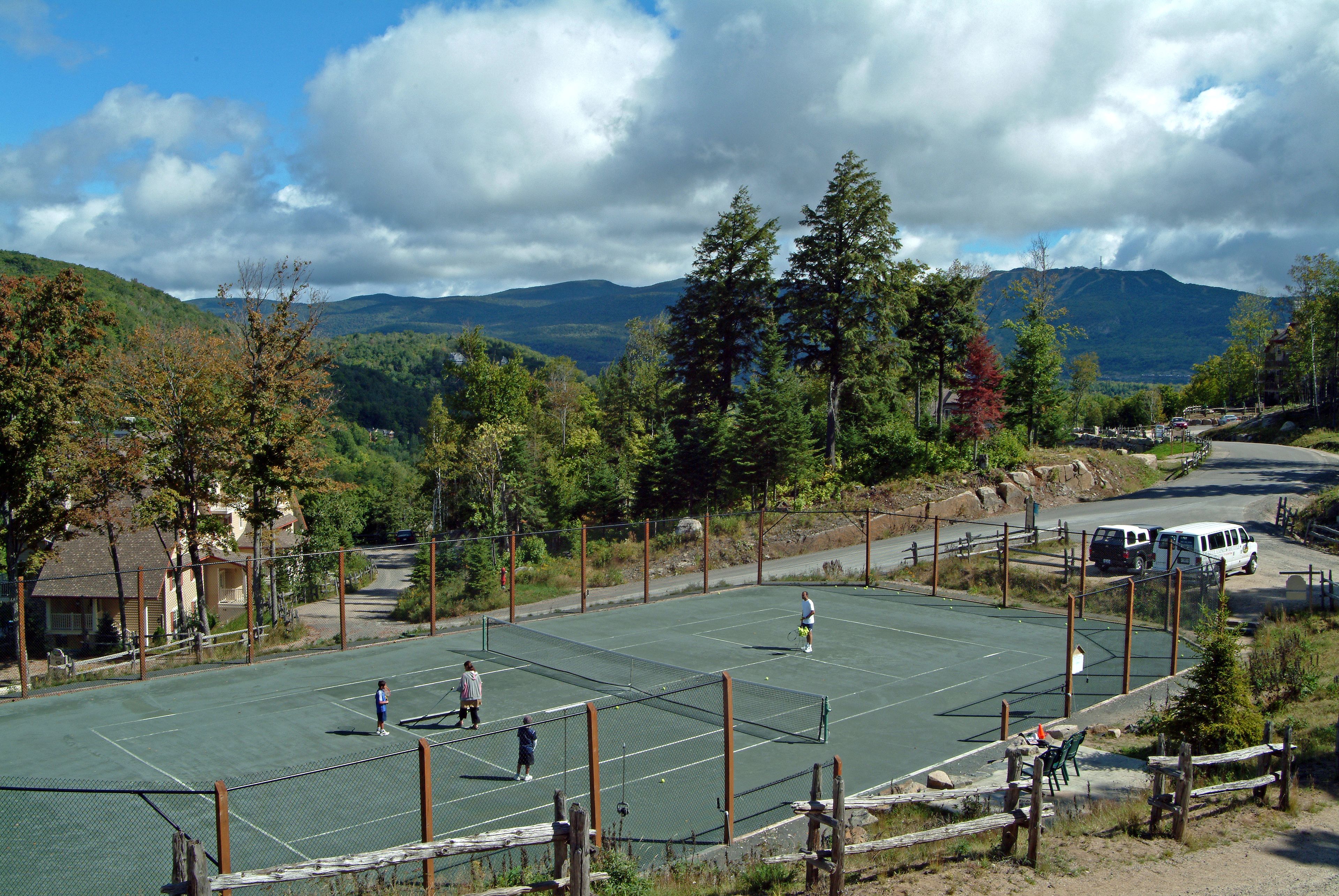 A tennis court surrounded by trees and mountains, with a few people playing.