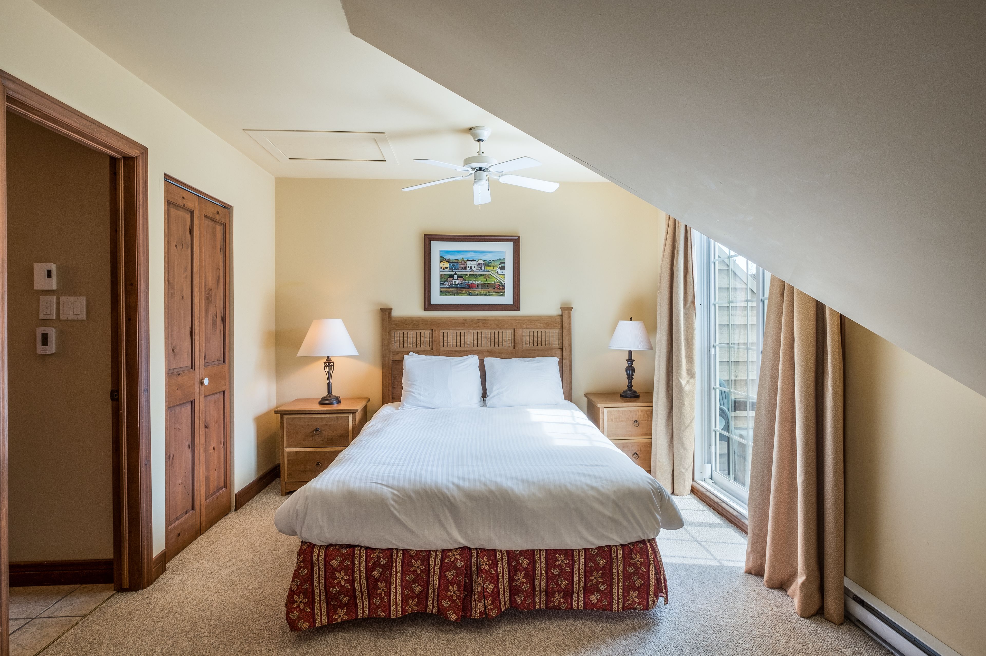 A hotel room with a large bed, wooden headboard, two bedside lamps, a ceiling fan, and a window with curtains.