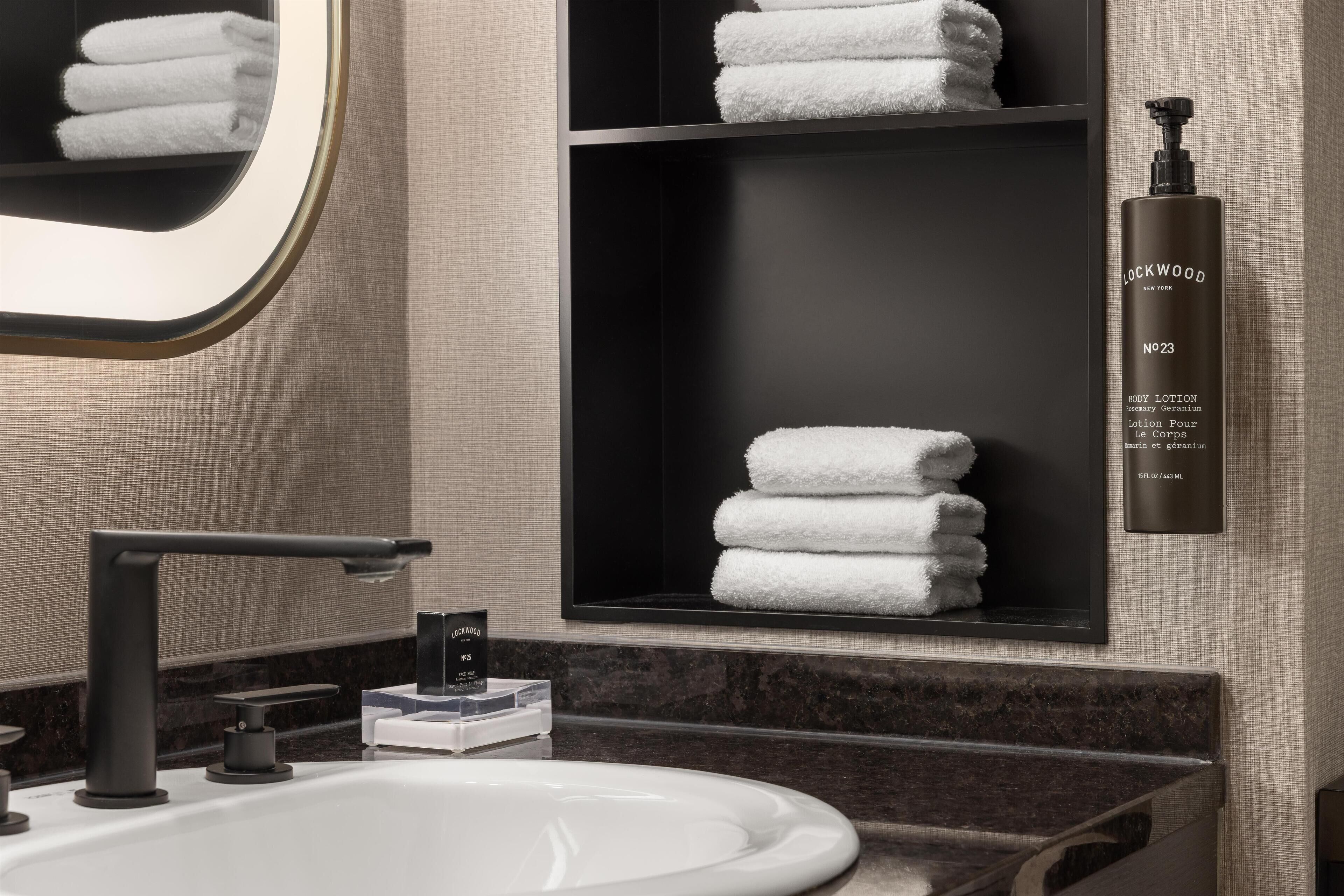A modern bathroom with a large mirror, a black shelf with folded towels, a white sink, and a bottle of Lockwood body lotion.