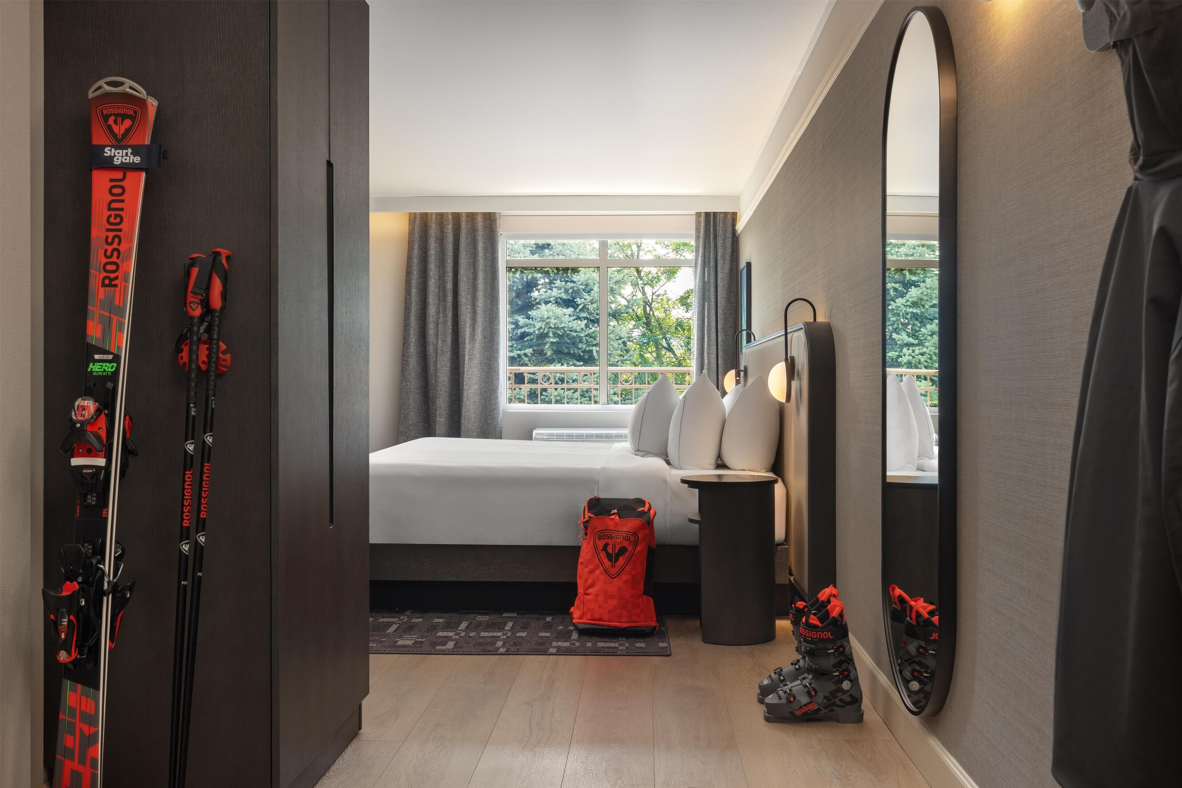A hotel room with a bed, a large mirror, and skis leaning against the wall.