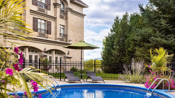 A swimming pool with lounge chairs and an umbrella, surrounded by a fence and greenery. - Hotel Vallea Bromont, Tapestry Collection by Hilton (Bromont)