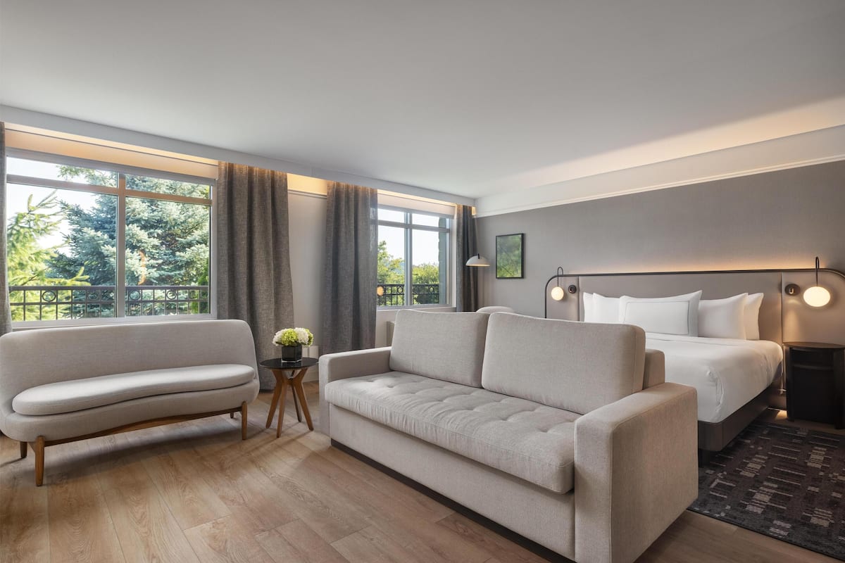 A modern hotel room with a sofa, a bed, a nightstand, and a large window with curtains.