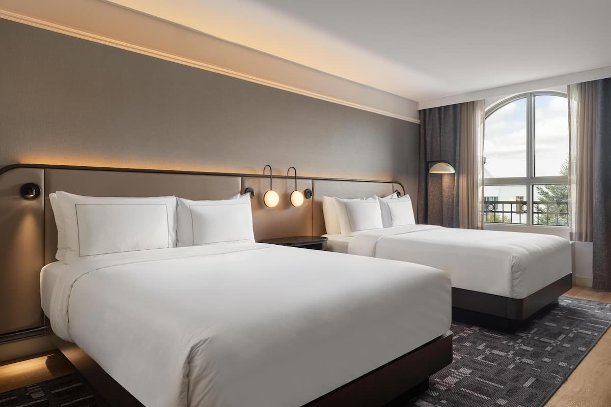 A hotel room with two beds, a large window with curtains, and a modern headboard with built-in lighting.