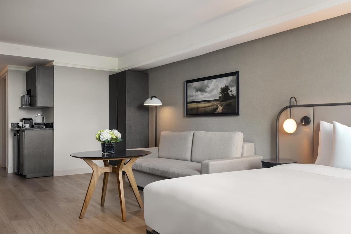 A modern hotel room with a bed, a sofa, a small table with flowers, a lamp, a painting on the wall, and a kitchenette in the background.