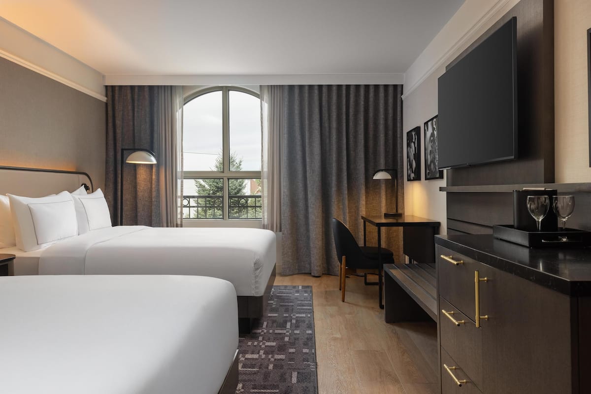 A hotel room with two beds, a desk, a television, and a large window with curtains.