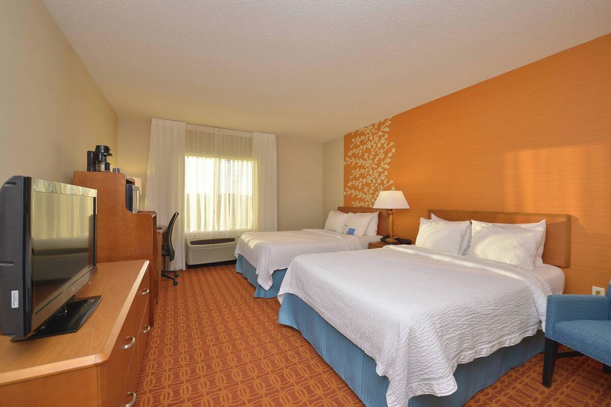 A hotel room with two beds, a desk with a television, a lamp, and a window with curtains.