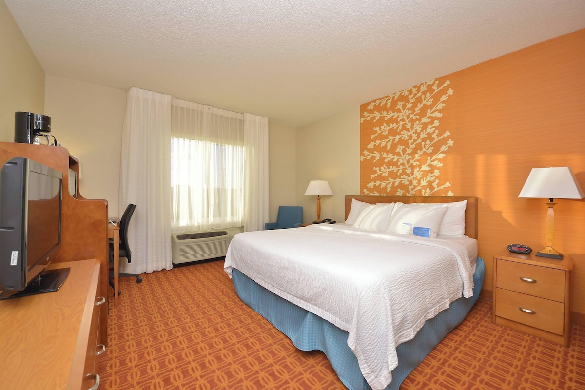 A hotel room with a bed, a desk with a computer, a chair, a lamp, and a wall decoration.