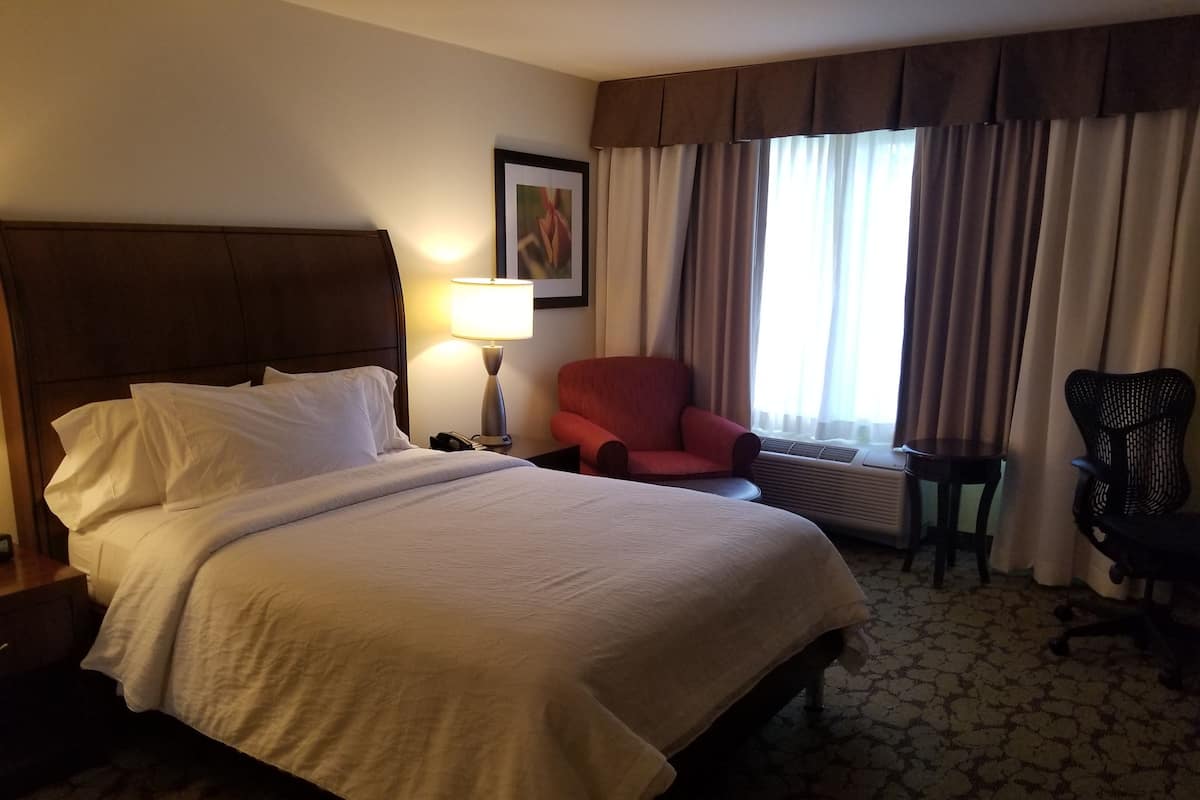 Une chambre d’hôtel avec un grand lit, un fauteuil rouge, un bureau avec une lampe et une fenêtre avec des rideaux.