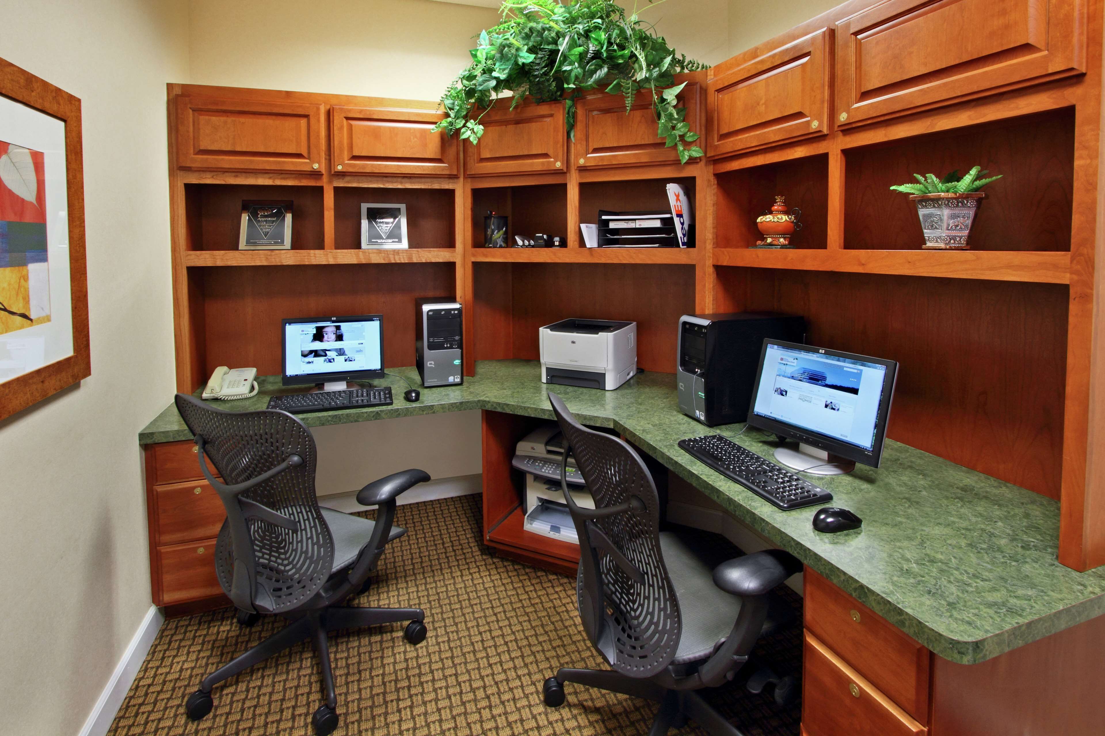 A home office with two desks, each with a computer, keyboard, and chair. There are shelves with decorative items and a potted plant.