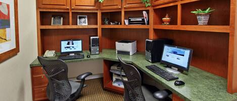 A home office with two desks, each with a computer, keyboard, and chair. There are shelves with decorative items and a potted plant.