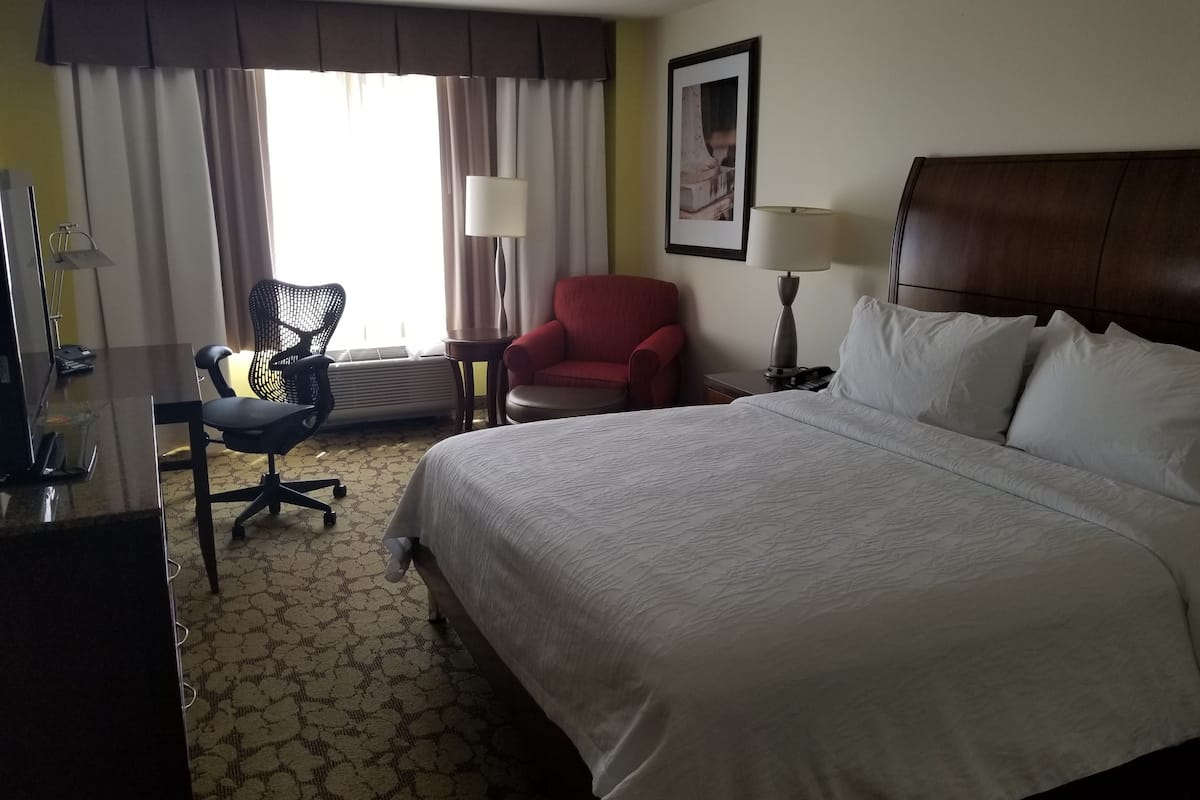 Une chambre d’hôtel avec un grand lit, un bureau avec une chaise, un fauteuil rouge et une fenêtre avec des rideaux.